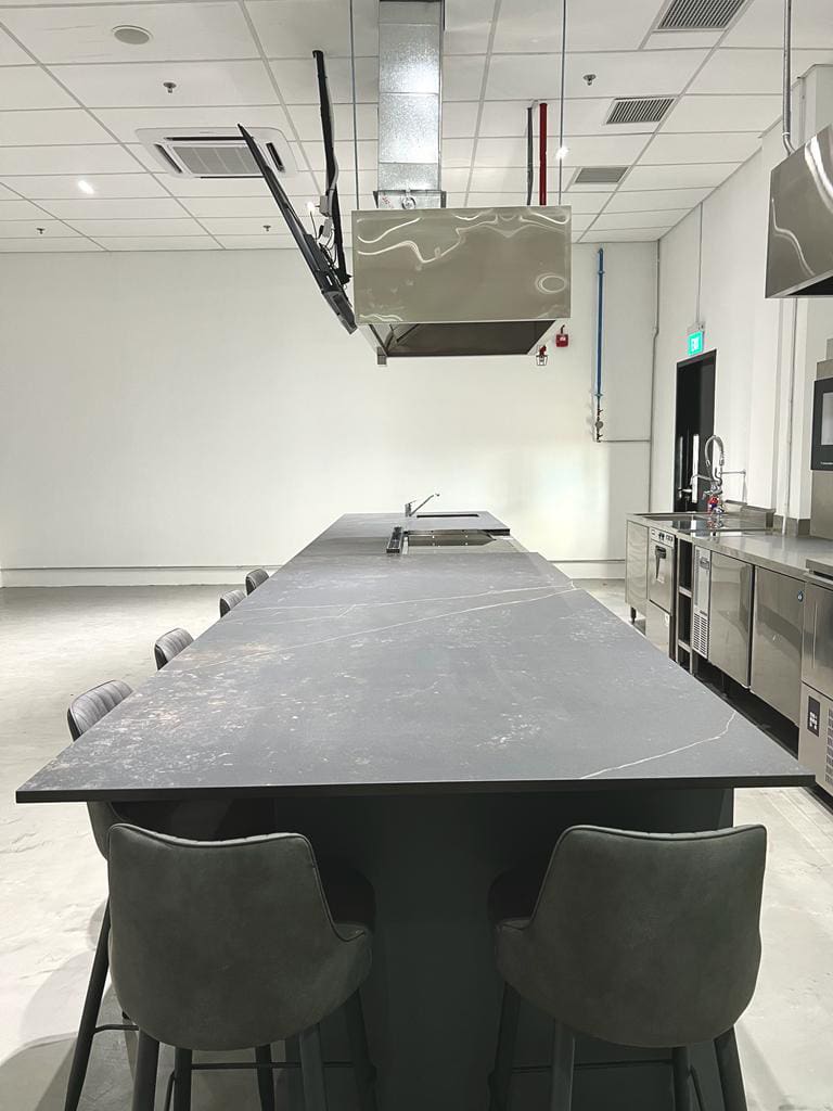 leinteri-gourmet-partner-partition-open-desks Commercial kitchen island counter featuring dark stone slab, high bar stools, and stainless steel exhaust hood